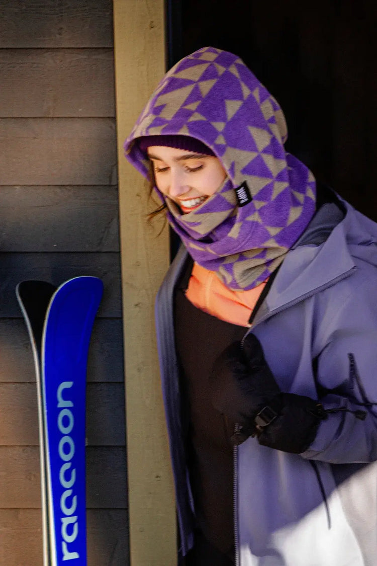 femme souriant à côté d'une paire de ski et portant une capuche nopé de couleur étoile mauve