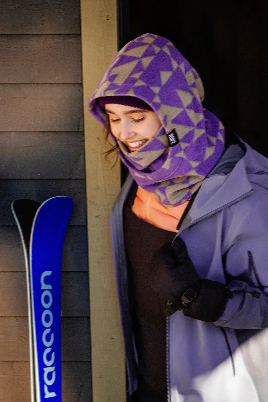 femme souriant à côté d'une paire de ski et portant une capuche nopé de couleur étoile mauve